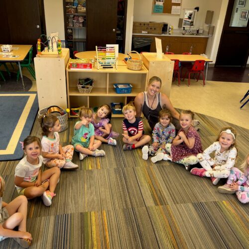 group of preschoolers in a half circle sitting on the floor with their teacher smiling at the camera