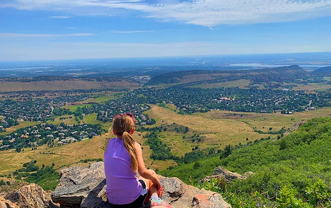 Person sitting on a rocky overlook, looking out over a wide valley with neighborhoods, open fields, and distant hills under a blue sky.